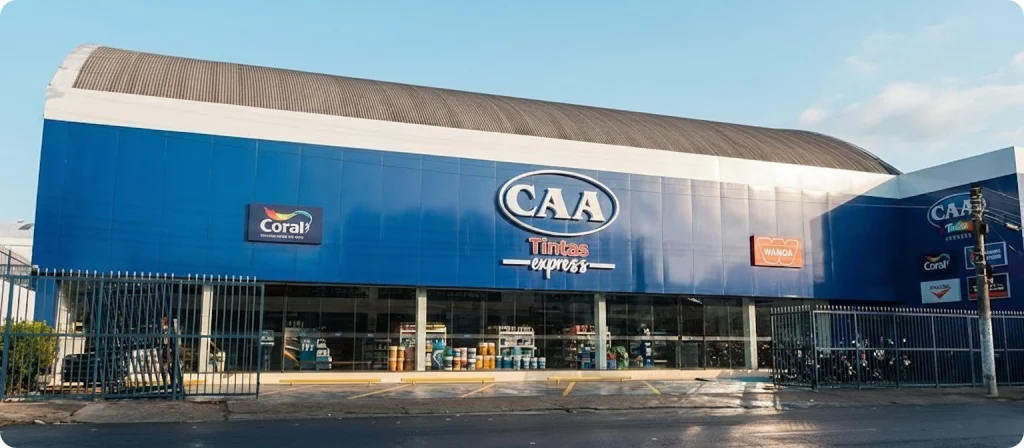 Fotografia profissional e cinematográfica em ângulo lateral baixo da fachada da loja CAA Tintas Express. O prédio possui uma arquitetura moderna e imponente, com uma ampla testeira em azul-marinho profundo onde se destaca o logotipo oval da "CAA Tintas Express" ao centro. A fachada é limpa e bem iluminada, exibindo também logotipos das marcas parceiras Coral e Wanda. Na parte inferior, uma grande vitrine de vidro permite ver o interior organizado da loja. O teto metálico curvo é visível sem obstruções, e o céu ao fundo está em um tom de azul claro e limpo, transmitindo uma sensação de clareza e profissionalismo.