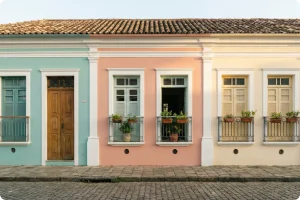 Fotografia profissional de uma fachada de estilo clássico e europeu, possivelmente uma confeitaria, com pintura em tons pastéis seguindo uma paleta harmoniosa de azul-claro, creme e detalhes em rosa suave e dourado. À esquerda, uma grande vitrine de vidro exibe doces e bolos refinados em suportes de cerâmica. No centro, uma porta de madeira azul-claro com uma placa onde se lê "L'Aube Sucrée". A fachada é adornada com trepadeiras de flores lilás (glicínias) e roseiras com rosas cor-de-rosa claro que emolduram a entrada. A iluminação é suave e natural, transmitindo uma sensação de leveza, elegância e delicadeza.